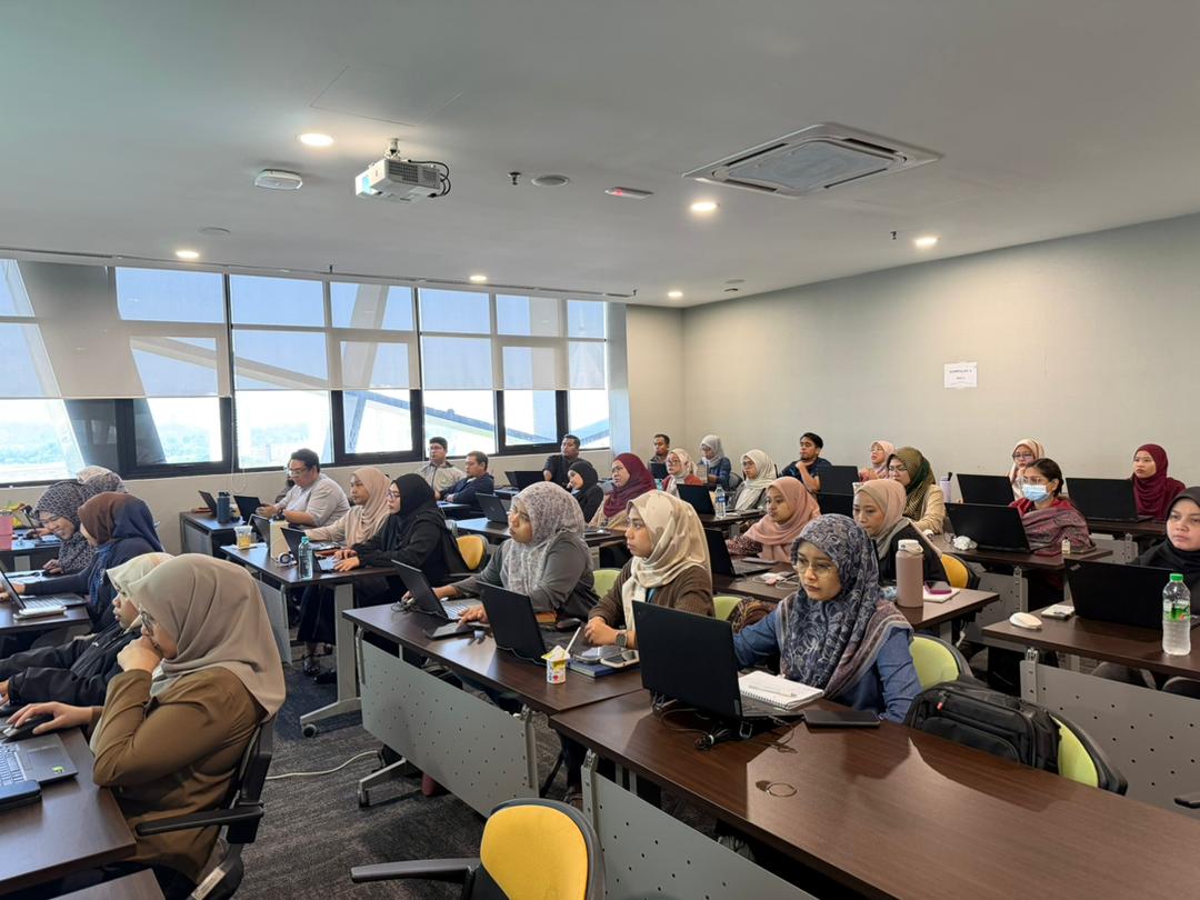 Classroom-style training session with attendees working on laptops at long desks beside large windows.